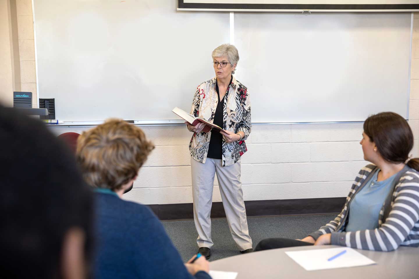 A professor with an open book in her hands reading to a class of graduate students