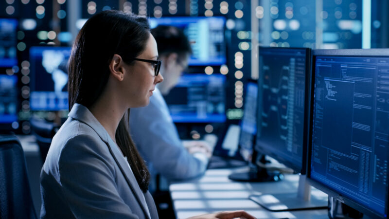 Female Engineer Controller Observes Working of the System. In the Background People Working and Monitors Show Various Information. Cybersecurity